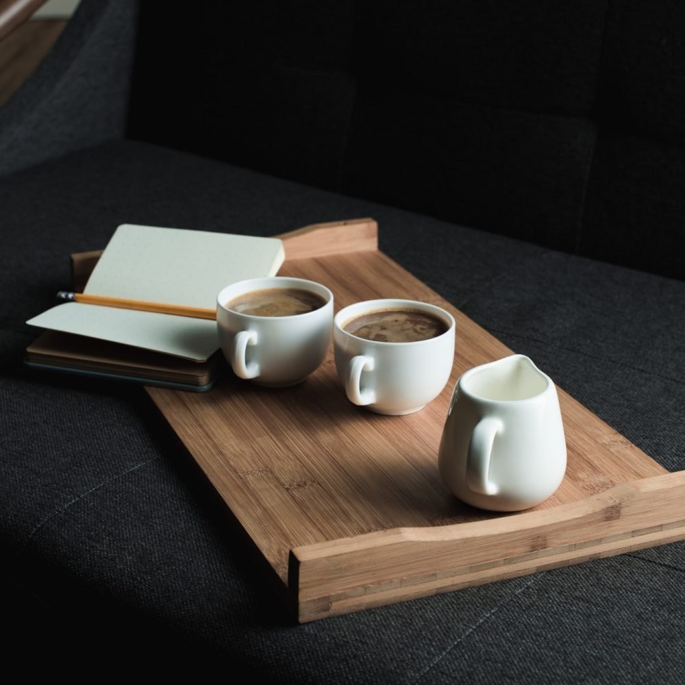 two cups of coffee jug of milk and notebook with pencil on wooden tray on sofa